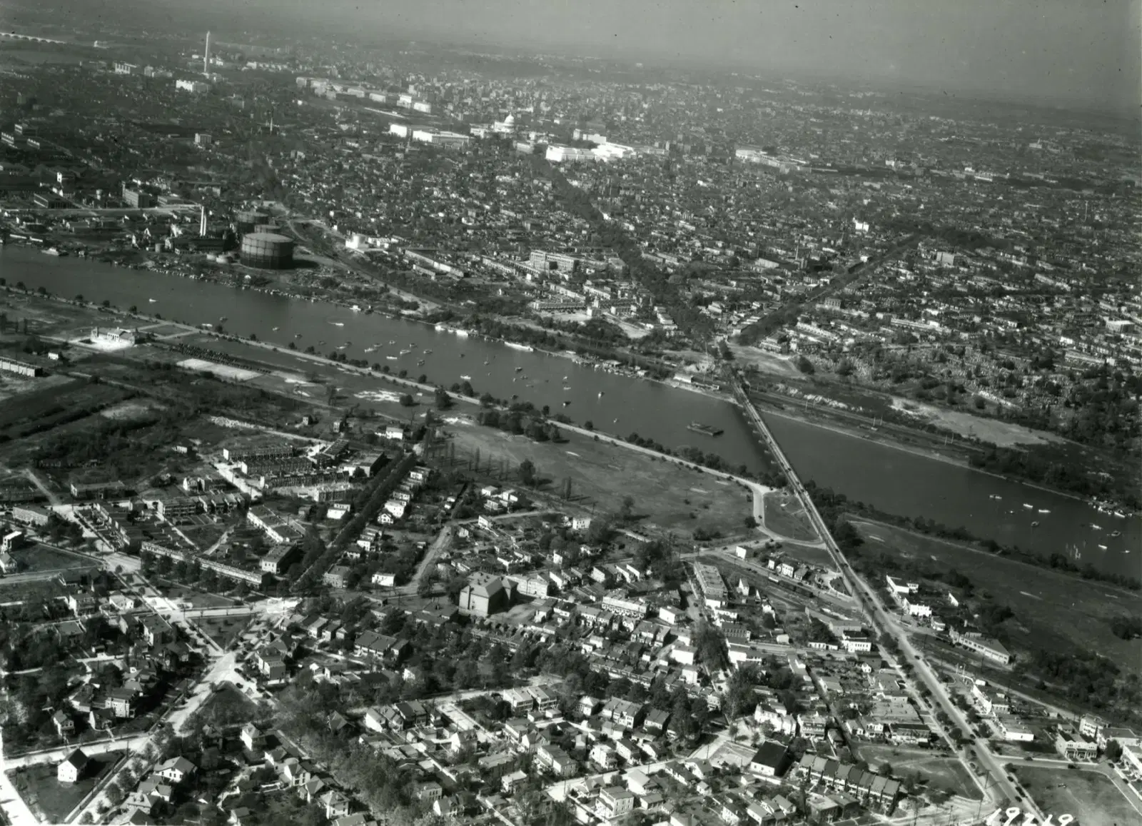 A 1932 aerial over the Anacostia River — small boats scattered across the water, the gasworks holder on the far bank, the Pennsylvania Avenue bridge, and the Capitol dome and Washington Monument on the skyline beyond.