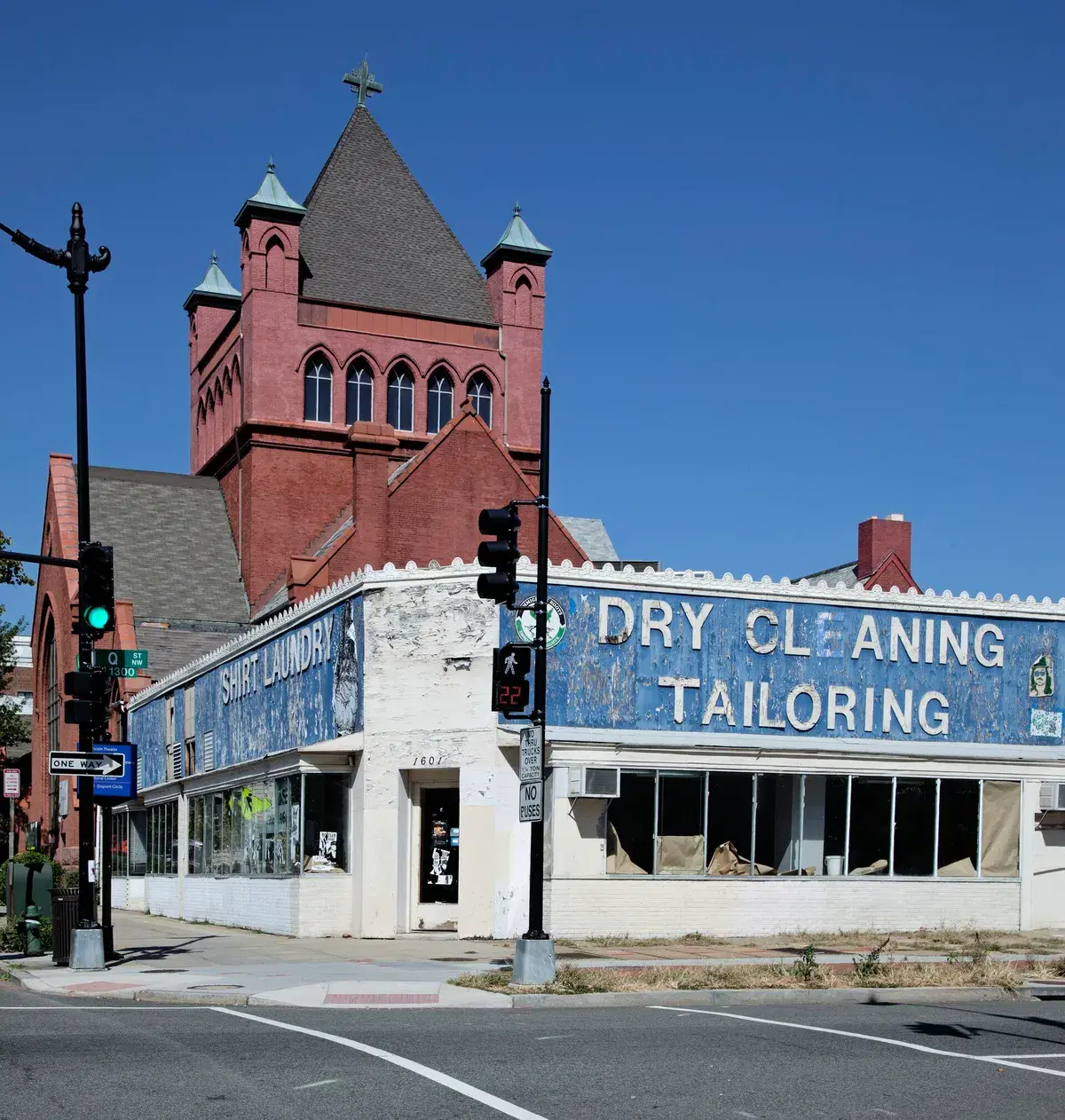 A one-story white corner building with faded blue Dry Cleaning and Tailoring signs, a red brick church tower behind it