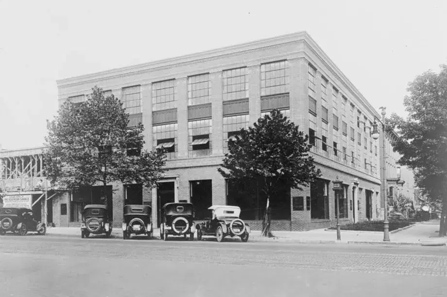 A three-story brick car showroom on a corner with four early automobiles parked in front