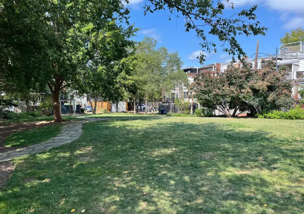 Green lawn and stone path of Crispus Attucks Park in summer, ringed by Bloomingdale rowhouse backs