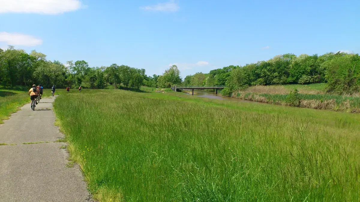 Cyclists on the paved Oxon Run trail beside the stream, meadow, and a footbridge, Washington, D.C.