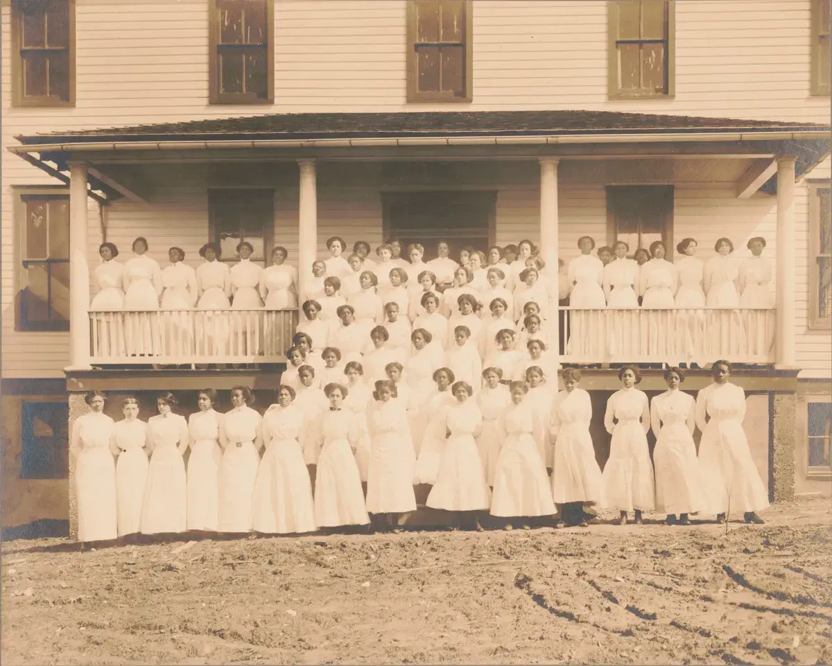 More than seventy young women in white dresses on the porch and front yard of the Training School's frame building
