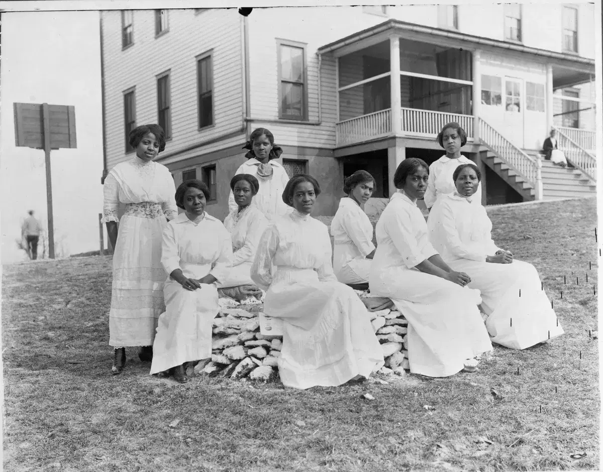 Nine young women in white dresses seated on a stone wall in front of the Training School's frame buildings