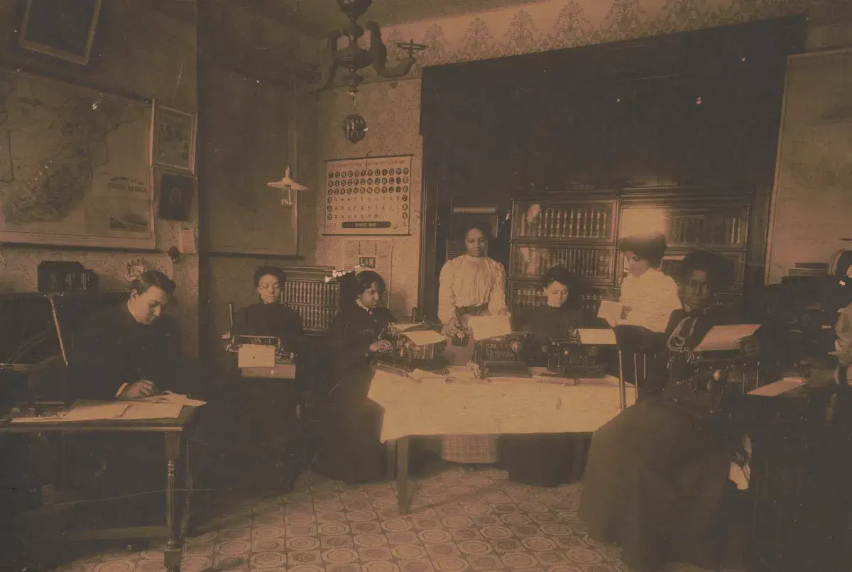 Young women at typewriters around a table in a Training School classroom, with maps and a keyboard chart on the walls