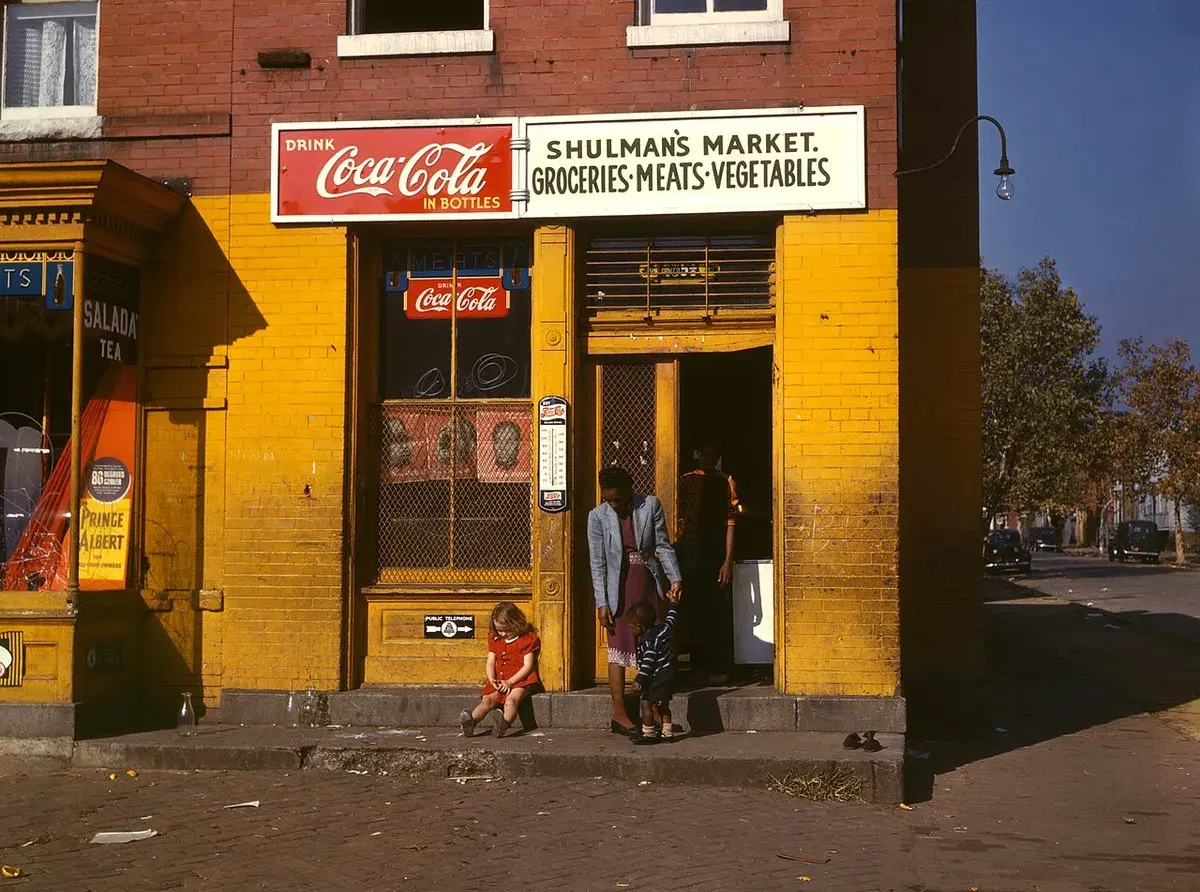 A woman and two children on the step of Shulman's Market, a yellow-painted corner grocery with a Coca-Cola sign