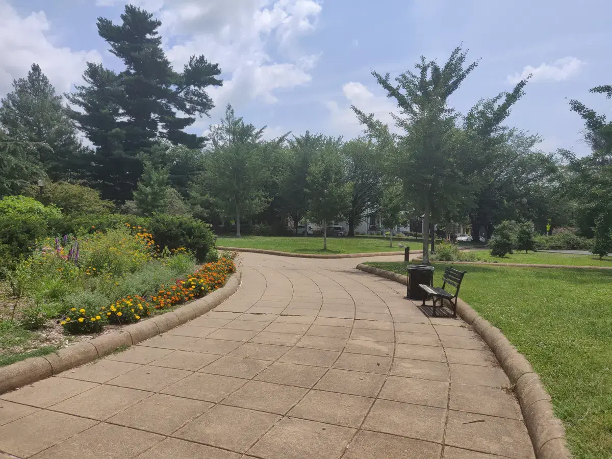 Summer park view inside Grant Circle: curving paved path, flower beds, bench, lawns with Petworth houses beyond