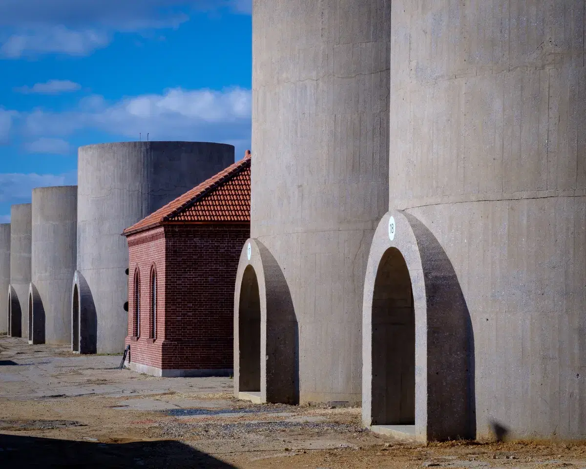 A row of tall concrete sand bins with arched doorways and a red-brick regulator house with a tile roof
