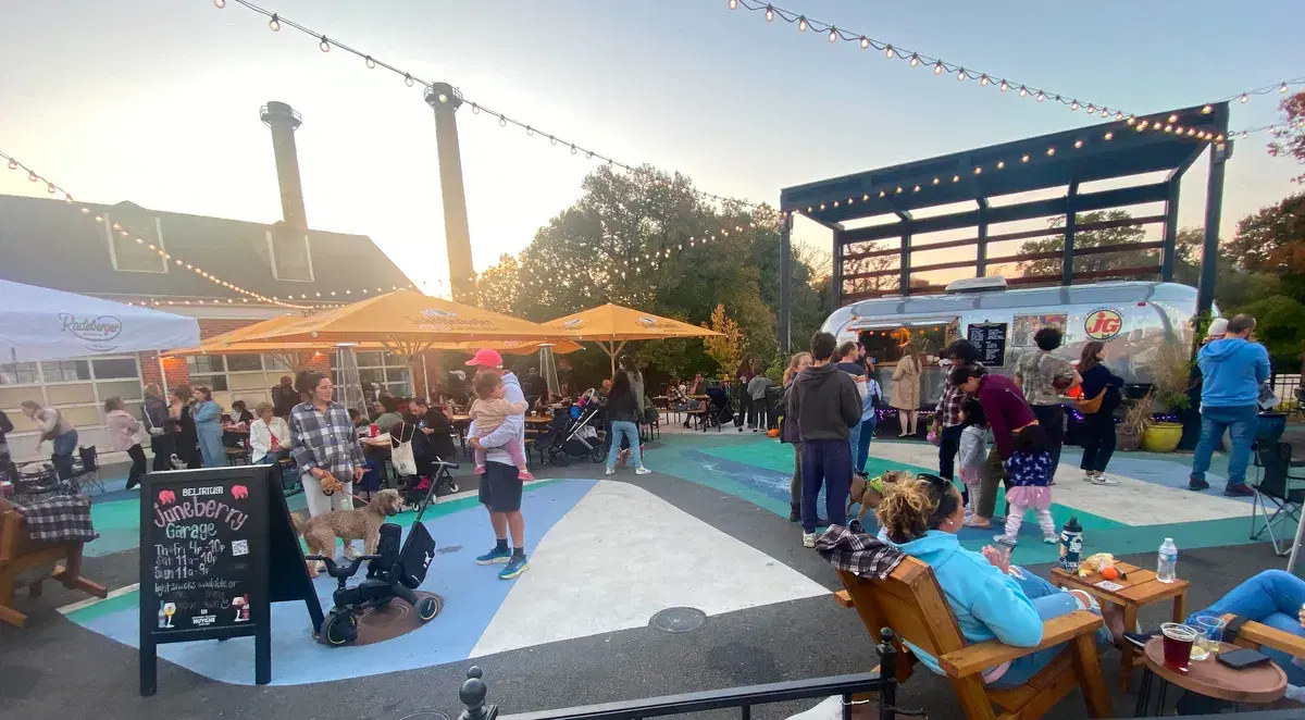 Juneberry Garage's busy patio under string lights and umbrellas, with the smokestacks behind