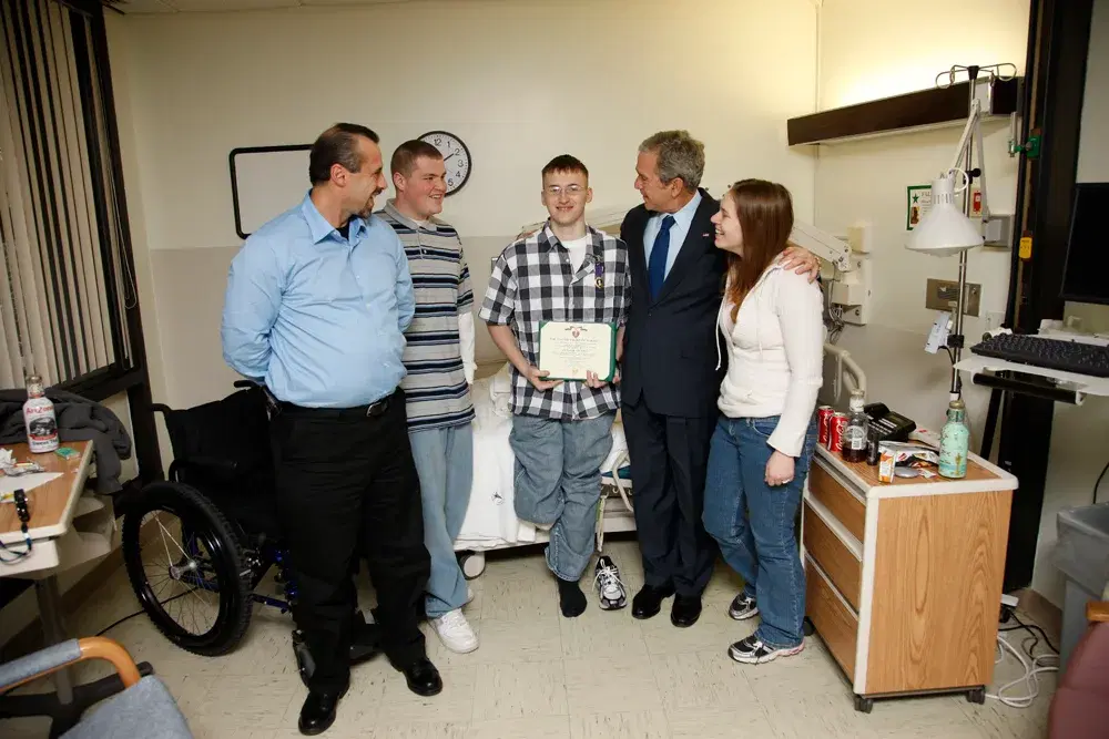 President George W. Bush with a soldier holding his Purple Heart certificate, the soldier's family and his hospital room behind them