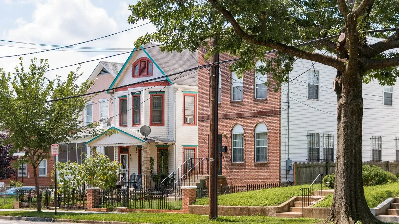 A tree-lined residential street in Congress Heights, Washington, D.C., with a Victorian frame house, sunflowers in the front garden, and brick rowhouses alongside.