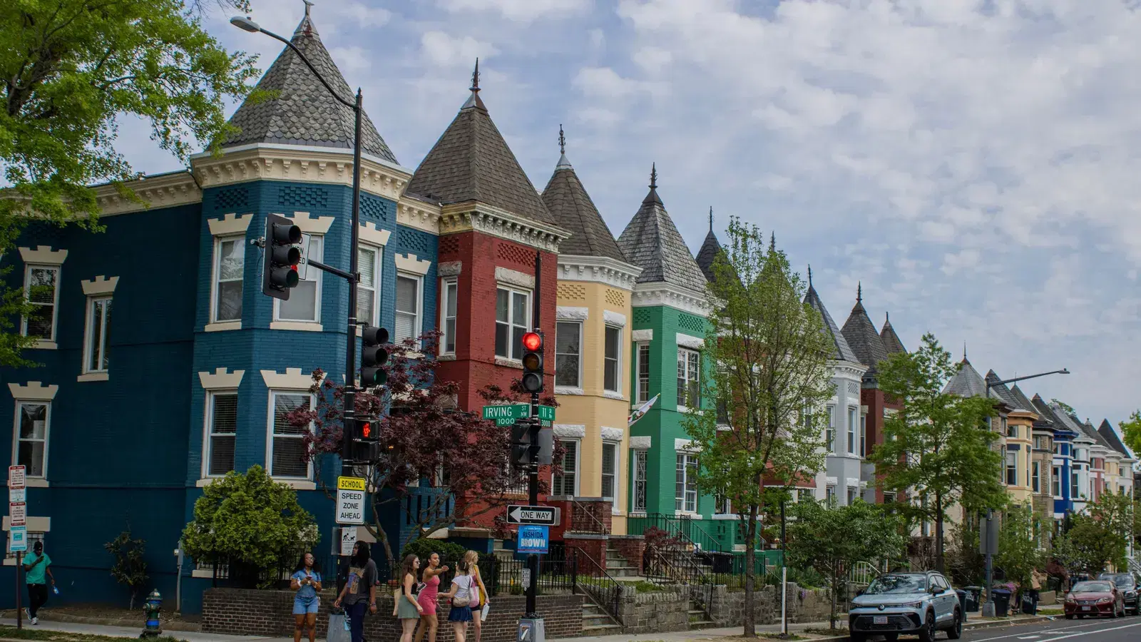 A corner of brightly painted, turreted Victorian rowhouses on a sunny Washington, D.C. street, with pedestrians crossing in the foreground.