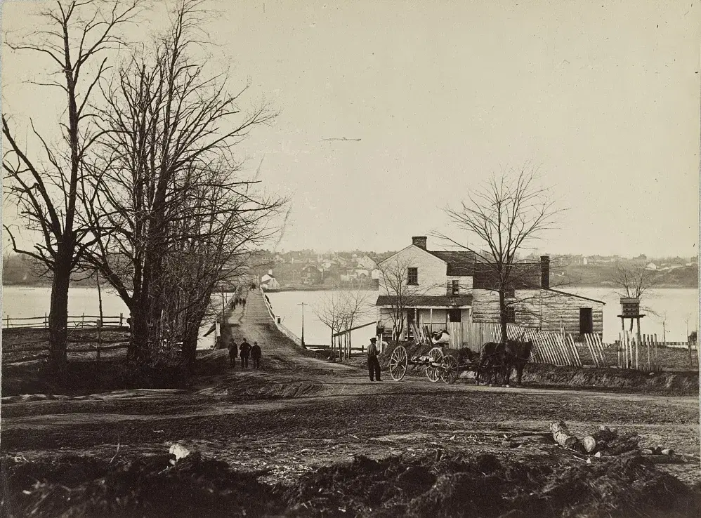 The long wooden Navy Yard bridge across the Eastern Branch of the Potomac in April 1865, seen from the east bank, with a horse and buggy waiting beside the toll house.