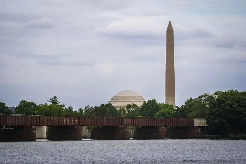 Real DC: Washington Monument and Jefferson Memorial dome seen across the water past a rail bridge - honest across-the-river DC vista matching the card's monuments-across-the-river theme