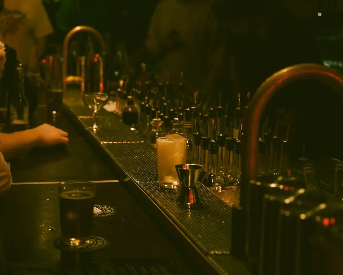 A moody, dimly lit cocktail bar rail lined with bottles and a finished cocktail.