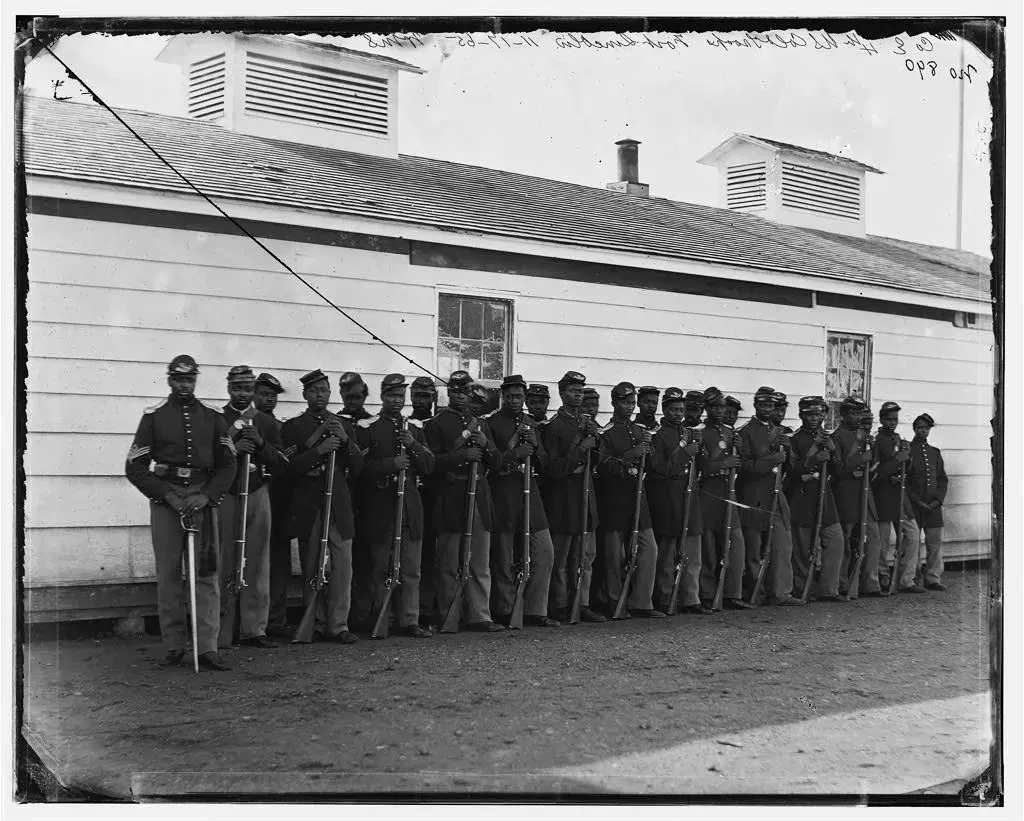 Soldiers of Company E, 4th U.S. Colored Infantry, standing in a line with rifles in front of a white wooden barracks