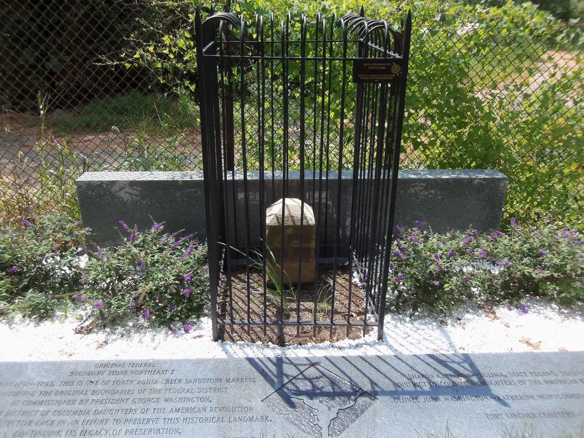 A weathered sandstone boundary marker inside a black iron cage, with a stone plaque in front
