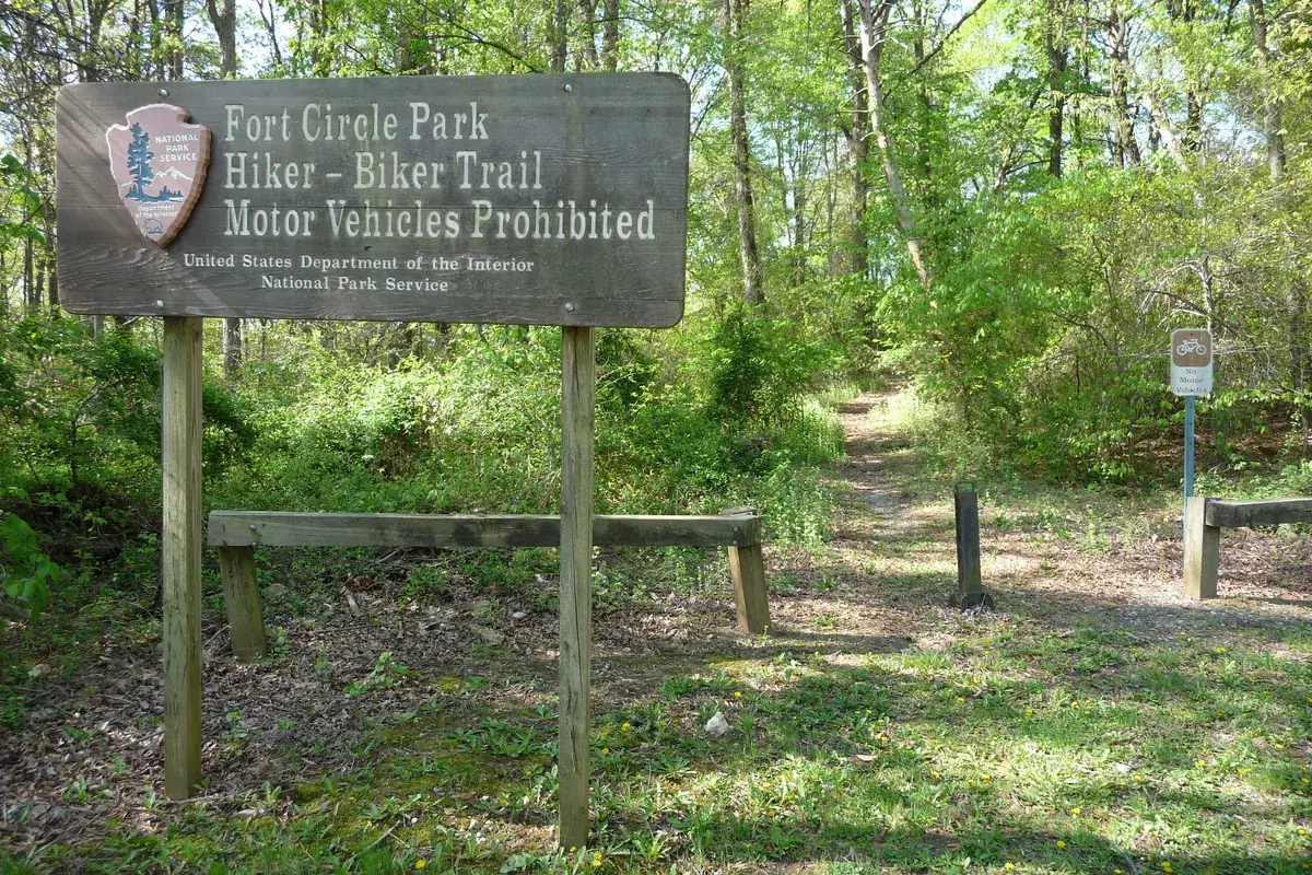 A wooden National Park Service sign for the Fort Circle Park Hiker-Biker Trail beside a path into the woods