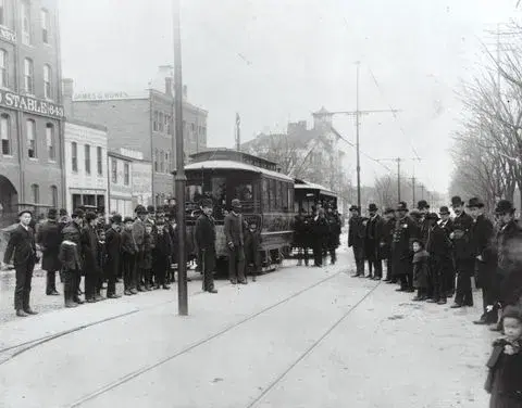 A crowd of men in hats standing around two electric streetcars on a city street