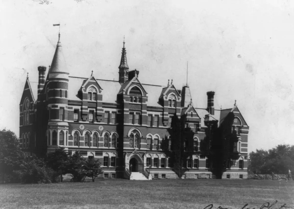 A large Victorian Gothic college building with towers and striped brickwork behind a lawn