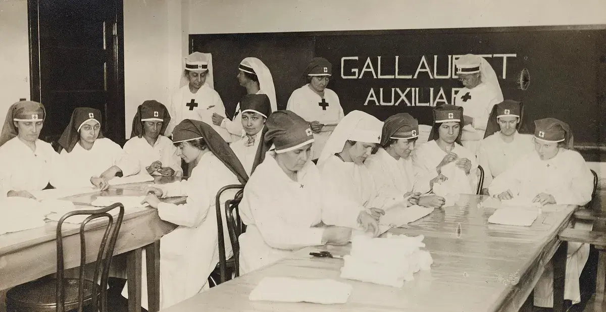 Women in Red Cross uniforms folding dressings at long tables in front of a Gallaudet Auxiliary sign