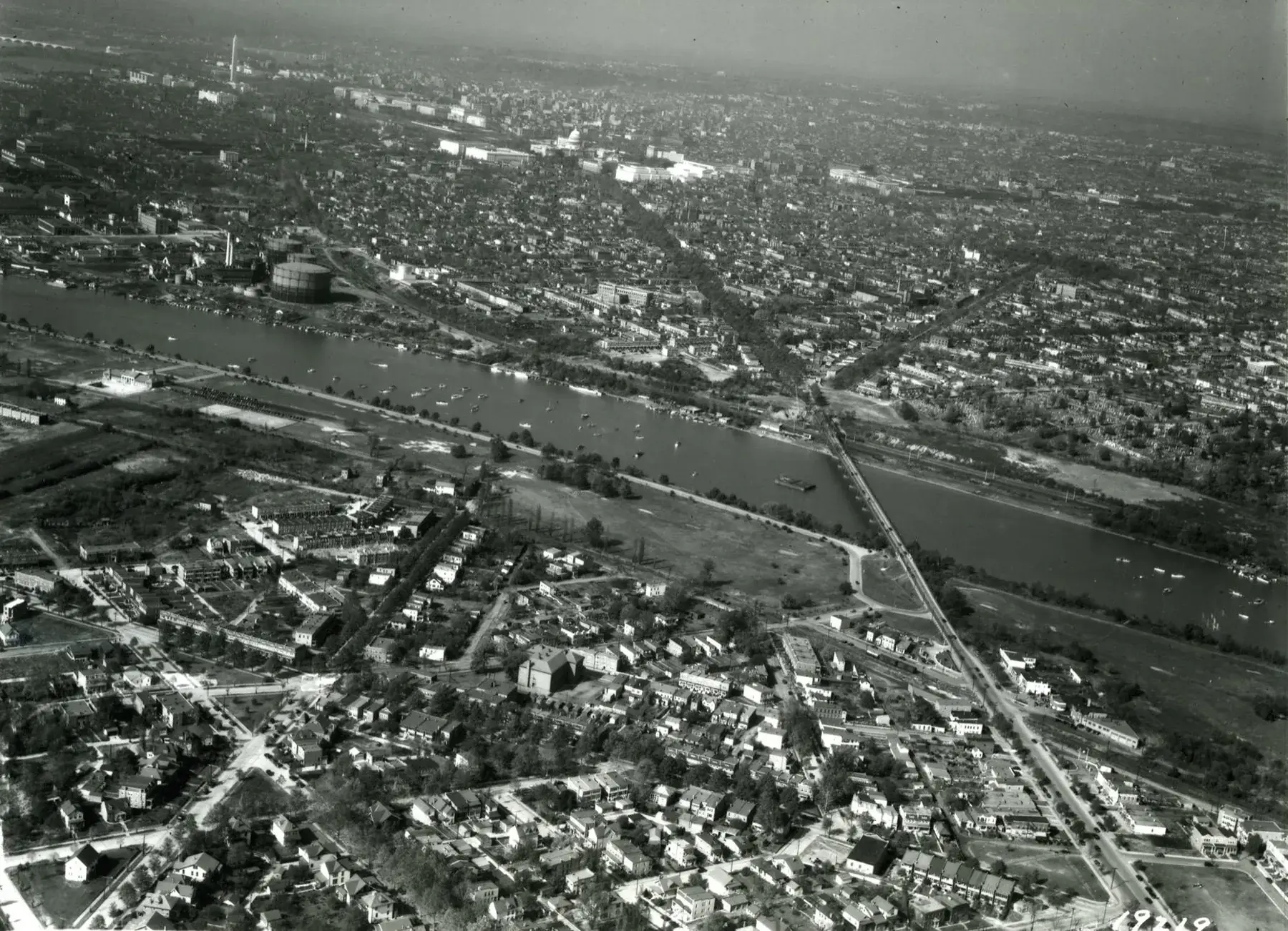 A 1932 aerial over the Anacostia River — small boats scattered across the water, the gasworks holder on the far bank, the Pennsylvania Avenue bridge, and the Capitol dome and Washington Monument on the skyline beyond.