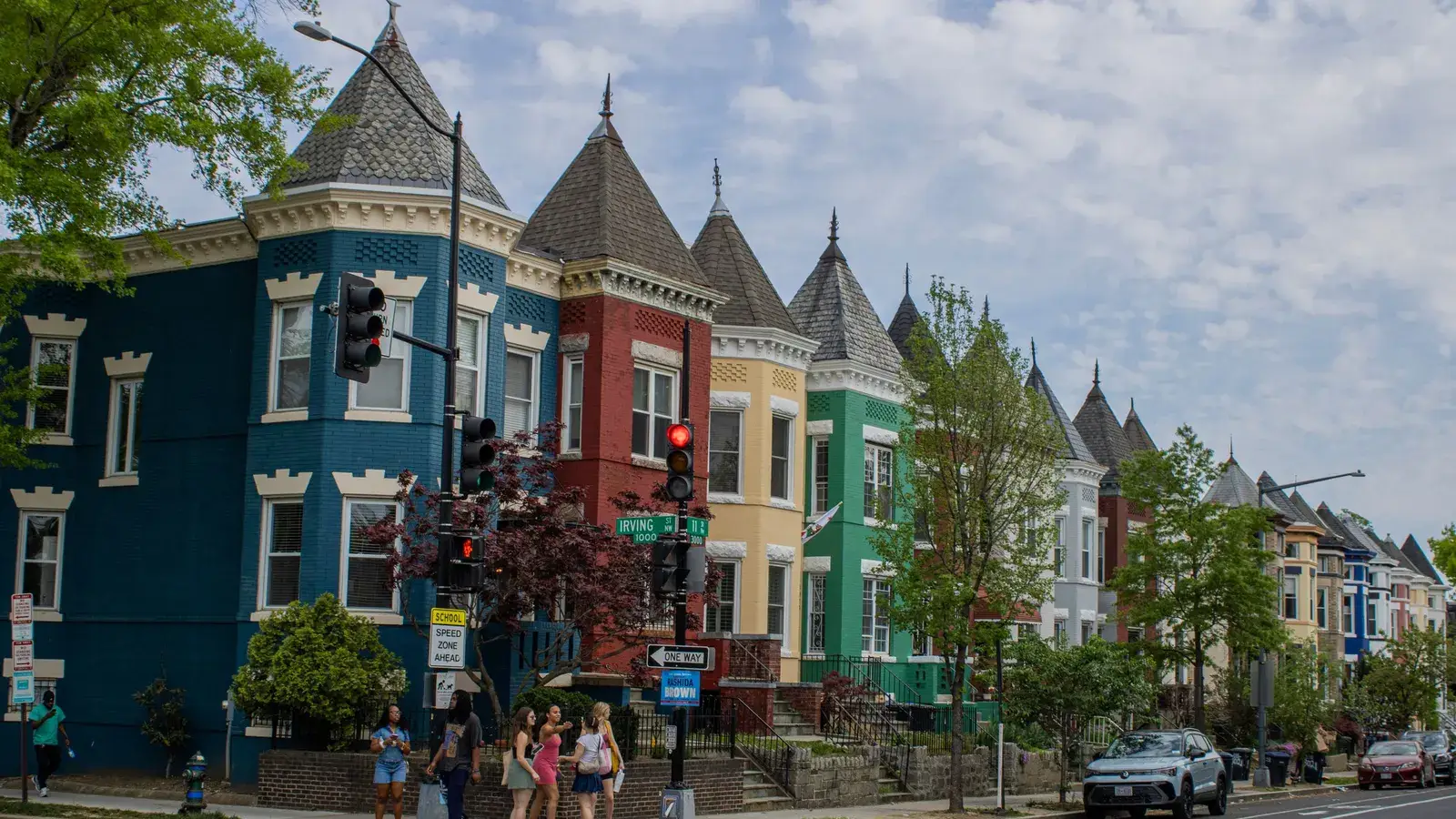 A corner of brightly painted, turreted Victorian rowhouses on a sunny Washington, D.C. street, with pedestrians crossing in the foreground.